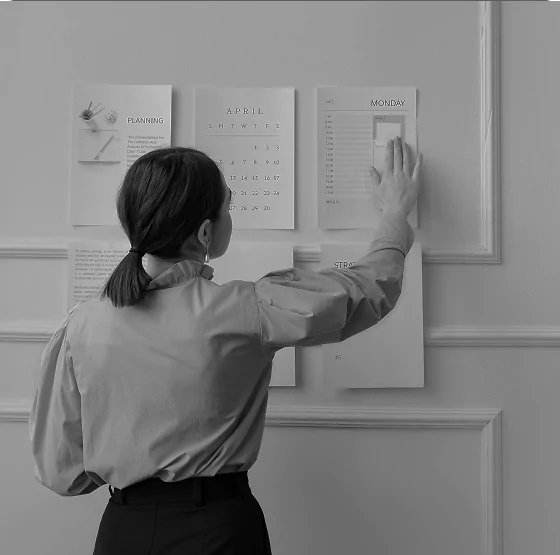 A woman standing in front of a wall with planning documents and a calendar, reaching toward a paper labeled "Monday," representing planning and scheduling tasks.