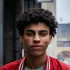 A young man with short curly hair wearing a red jacket, standing outdoors in an urban setting with a neutral expression.